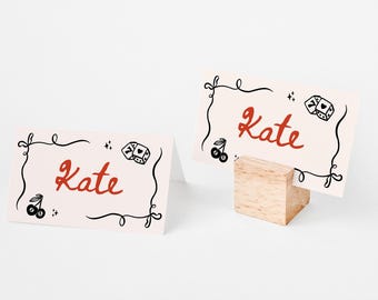 May include: Two white place cards with black and red designs. The cards have a black swirl design, a black cherry, and a black dice with red dots. The text "Kate" is written in red on each card.