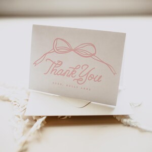 May include: A folded, light gray card with a pink bow illustration and the words "Thank You" in a cursive font. The text "xoxo, Holly Anne" is below the main text. The card is inside a white envelope.