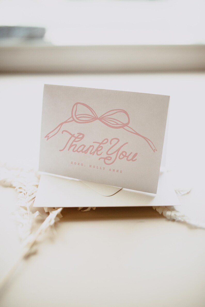 May include: A folded, light gray card with a pink bow illustration and the words "Thank You" in a cursive font. The text "xoxo, Holly Anne" is below the main text. The card is inside a white envelope.