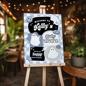 May include: A baby shower welcome sign on an easel. The sign has a blue and white checkered background with the words "Welcome to Kelly's Baby Shower". It features ghost and smiley face graphics. The sign also says "We're so happy you're here!"