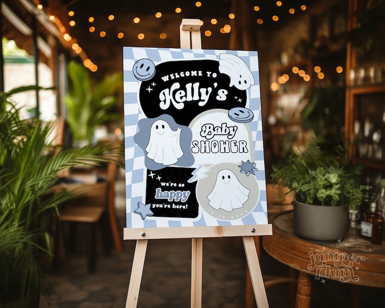 May include: A baby shower welcome sign on an easel. The sign has a blue and white checkered background with the words "Welcome to Kelly's Baby Shower". It features ghost and smiley face graphics. The sign also says "We're so happy you're here!"