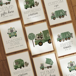May include: A set of framed prints featuring green garbage trucks and raccoons. The prints display phrases such as "Dump Gifts Here," "Refueling Station," and "Loading Zone." The signs are displayed on a wooden surface.