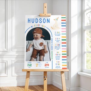 May include: A personalized first-year milestone poster on an easel. The poster features a photo of a baby wearing a baseball cap and a t-shirt. Text includes "HUDSON", "MY FIRST YEAR", and "I WEIGH 32 LBS, I HAVE 7 TEETH, I AM 29 INCHES".