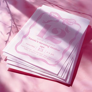 May include: A stack of pink and white birthday invitations. The invitations feature a large number 12, a bow, and the text "Annie's turning twelve!" with event details. The background is a soft pink.