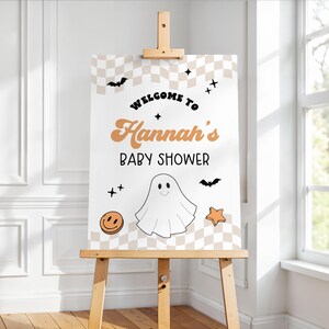 May include: A baby shower sign on a wooden easel. The sign features a checkered background with the words "Welcome to Hannah's Baby Shower" in orange and black. It includes a ghost, bats, stars, and a smiley face.
