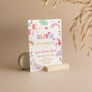 Puede incluir: Una invitaci&oacute;n colorida con un tema de unicornio para una fiesta de primer cumplea&ntilde;os. La invitaci&oacute;n presenta un arco&iacute;ris, estrellas, nubes y el nombre "Olivia" en una fuente caprichosa. El texto dice "Olivia cumple un a&ntilde;o! &iexcl;Ven a celebrar con nosotros! 15 de marzo de 2 a 4 pm Casa de Olivia 375 Juniper way, boulder, co RSVP a Cara al 123.456.7890"