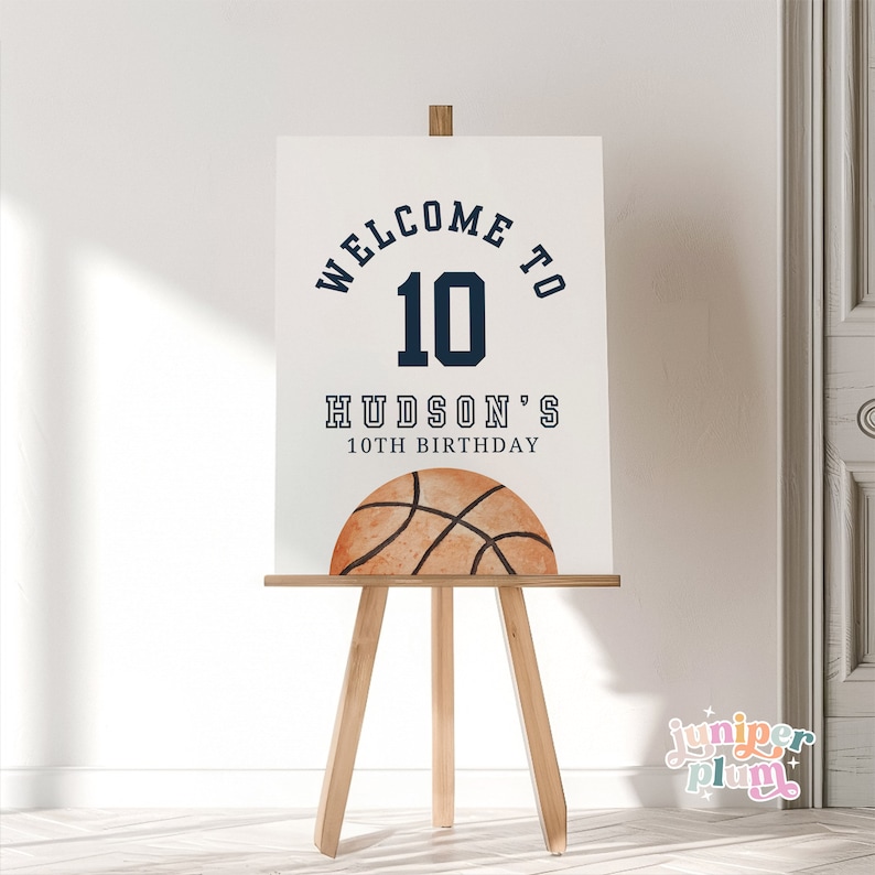 May include: White sign with blue text that says "Welcome to 10 Hudson's 10th Birthday". A brown basketball with black lines is at the bottom of the sign.
