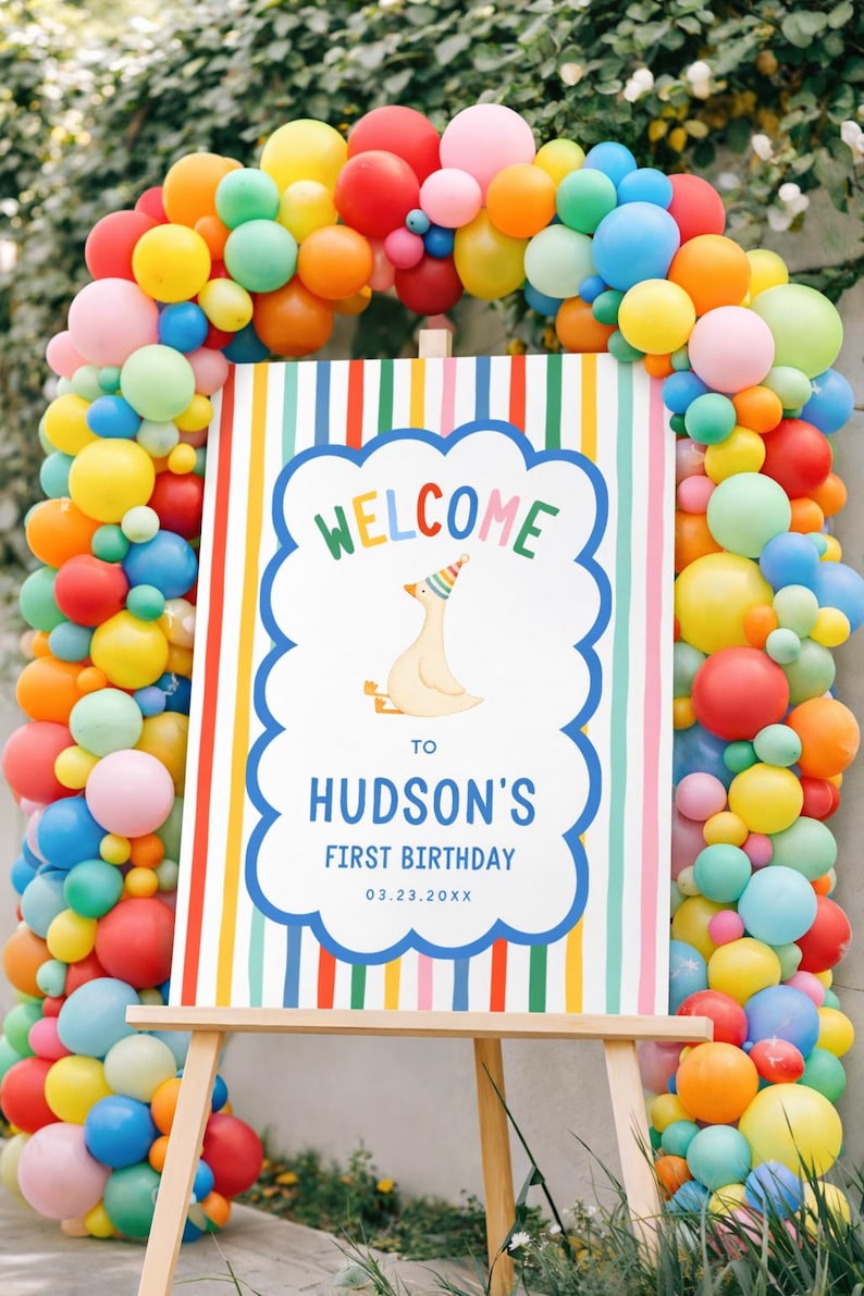 May include: A colorful birthday party sign on an easel, framed by a vibrant balloon arch. The sign reads "WELCOME to HUDSON'S FIRST BIRTHDAY 03.23.20XX" with a cartoon goose wearing a party hat. The balloons are in shades of red, yellow, blue, orange, and pink.