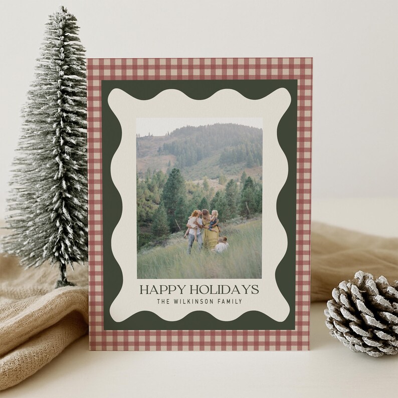 May include: A holiday card with a family photo set against a mountain backdrop. The card features a red and white gingham border, a dark green wavy frame, and the text "HAPPY HOLIDAYS" and "THE WILKINSON FAMILY". A small decorative Christmas tree and pinecone are in the background.