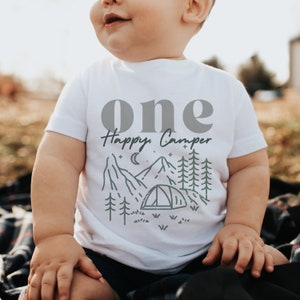 May include: White baby onesie with the text "one Happy Camper" in gray and black. The design features a mountain range, a tent, trees, and stars.