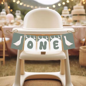 以下が含まれることがあります： 白と緑の旗に白い文字で「ONE」と書いてあり、両端に白いガチョウがいます。旗は白いハイチェアにかけられています。