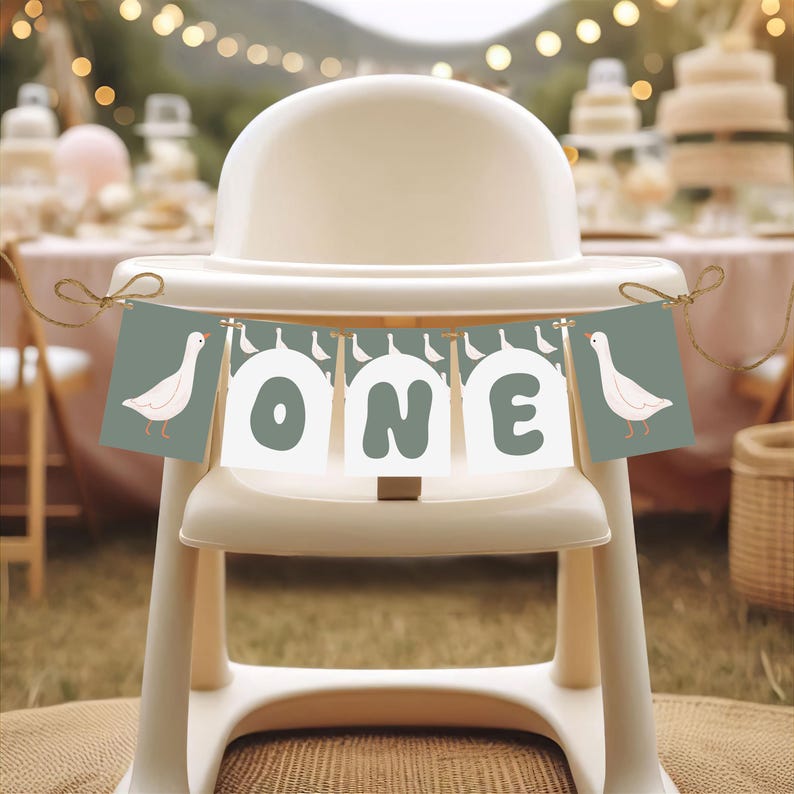 以下が含まれることがあります： 白と緑の旗に白い文字で「ONE」と書いてあり、両端に白いガチョウがいます。旗は白いハイチェアにかけられています。