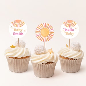Peut inclure: Trois cupcakes d&eacute;cor&eacute;s de gla&ccedil;age blanc et surmont&eacute;s d'une &eacute;toile dor&eacute;e et d'un rond de papier blanc. Les d&eacute;corations pr&eacute;sentent un soleil aquarelle avec des rayures roses et orange et un texte qui dit "Baby Smith", "Hello Baby" et un soleil avec des rayures roses et orange.