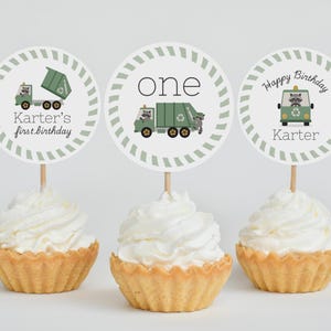 May include: Three cupcakes with white frosting and decorative toppers. The toppers feature a green garbage truck, the word "one", and a raccoon in a green vehicle. Text includes "Karter's first birthday" and "Happy Birthday Karter".