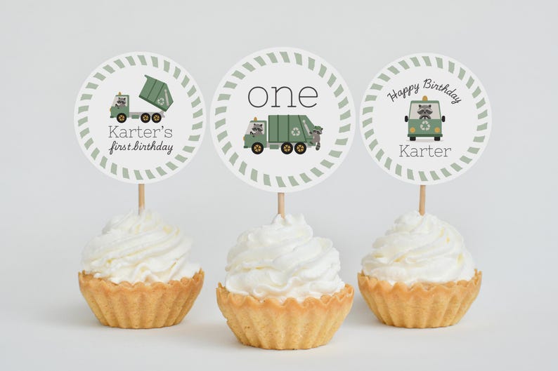 May include: Three cupcakes with white frosting and decorative toppers. The toppers feature a green garbage truck, the word "one", and a raccoon in a green vehicle. Text includes "Karter's first birthday" and "Happy Birthday Karter".