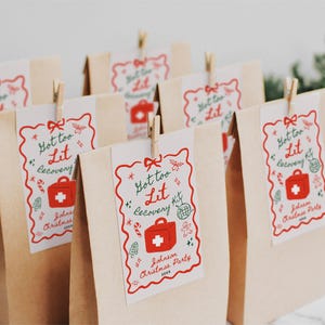 Puede incluir: Un conjunto de seis bolsas de papel marr&oacute;n con etiquetas rojas y verdes "Got too Lit Recovery Kit". Las etiquetas muestran un botiqu&iacute;n de primeros auxilios rojo con una cruz blanca y ramas de acebo verdes.