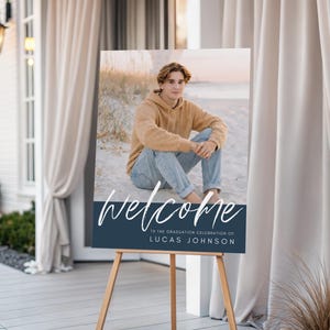May include: A graduation welcome sign on a wooden easel. The sign features a photo of a person in a tan hoodie and jeans. The word "welcome" is written in white script above a navy blue banner with the graduate's name.