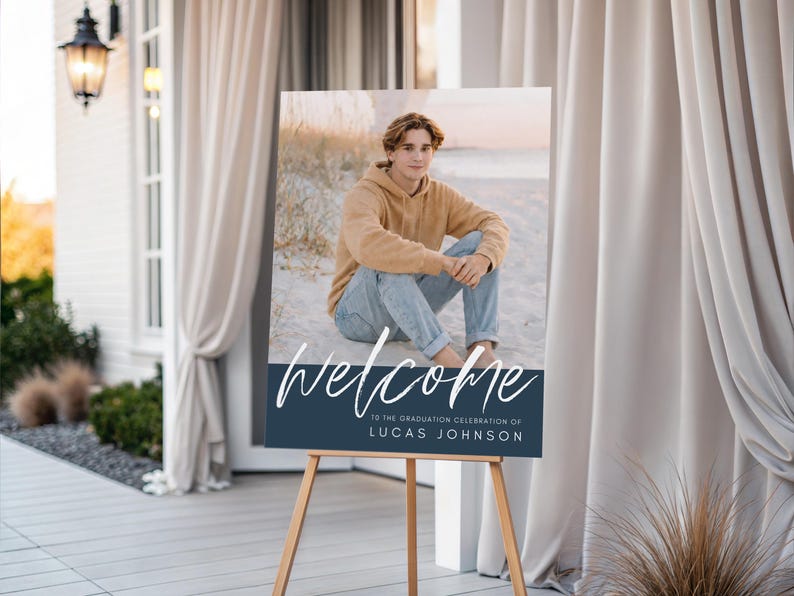 May include: A graduation welcome sign on a wooden easel. The sign features a photo of a person in a tan hoodie and jeans. The word "welcome" is written in white script above a navy blue banner with the graduate's name.