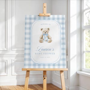 May include: A baby shower sign on a wooden easel. The sign features a light blue gingham pattern background with a white oval frame. Inside the frame is a teddy bear illustration with a blue bow, and the text "welcome to Lauren's BABY SHOWER".