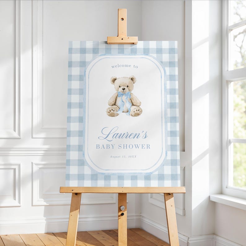 May include: A baby shower sign on a wooden easel. The sign features a light blue gingham pattern background with a white oval frame. Inside the frame is a teddy bear illustration with a blue bow, and the text "welcome to Lauren's BABY SHOWER".
