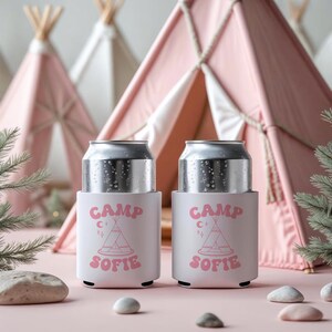 May include: Two silver beverage cans in white holders with the text "CAMP SOFIE" in pink. The holders feature a teepee graphic and are set against a pink background with miniature teepees.