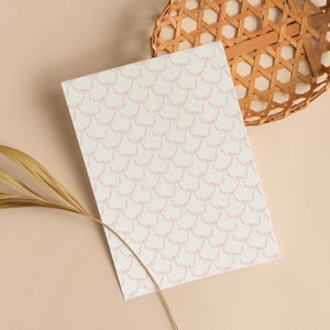 May include: A rectangular sheet of paper with a repeating pattern of light pink bows on a white background. A woven basket and a dried leaf are in the background, set against a beige surface.