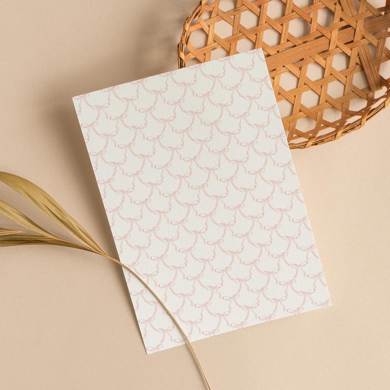 May include: A rectangular sheet of paper with a repeating pattern of light pink bows on a white background. A woven basket and a dried leaf are in the background, set against a beige surface.