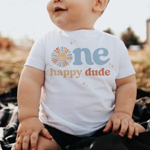 May include: White short-sleeved t-shirt with the text "one happy dude" in blue, orange, and yellow. The word "one" is above "happy dude" with a sun graphic. The shirt is for a baby.