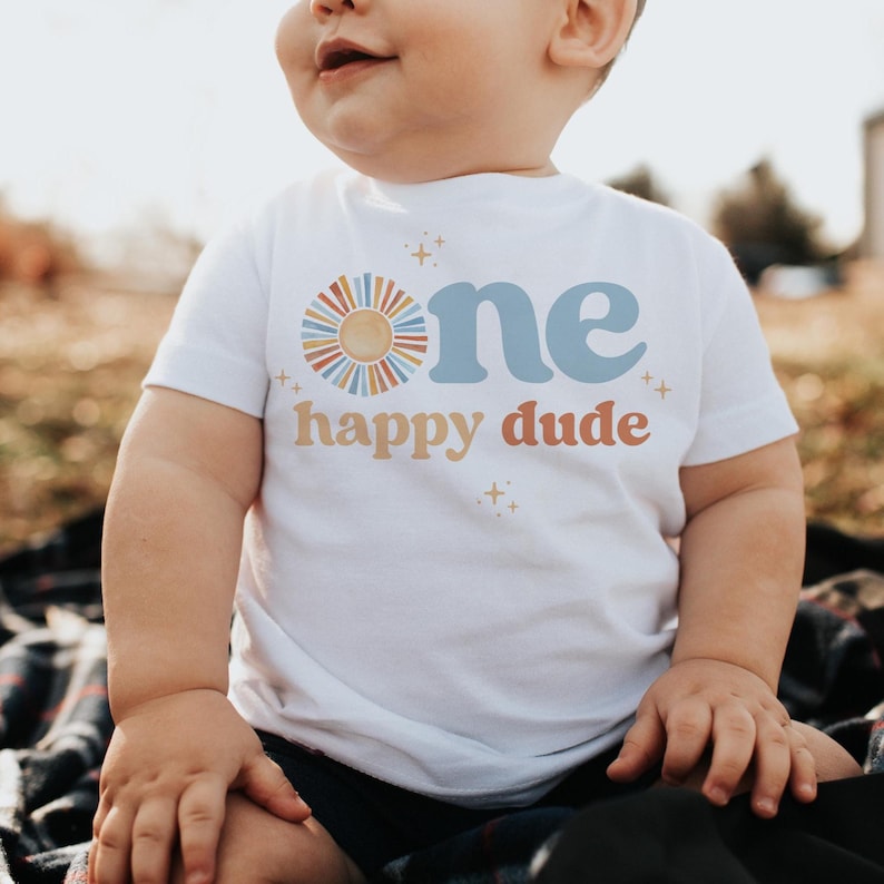 May include: White short-sleeved t-shirt with the text "one happy dude" in blue, orange, and yellow. The word "one" is above "happy dude" with a sun graphic. The shirt is for a baby.