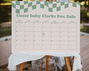 May include: A light beige calendar on a wooden easel with the text "Guess baby Clarks Due Date" in green. The calendar has a green checkered pattern at the top and a calendar grid with dates from March to April. A white cloth is draped below.