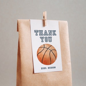 May include: A white thank you card with black text that says "THANK YOU" and a brown and orange basketball illustration. The card is attached to a brown paper bag with a wooden clothespin.
