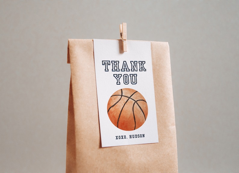 May include: A white thank you card with black text that says "THANK YOU" and a brown and orange basketball illustration. The card is attached to a brown paper bag with a wooden clothespin.