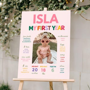 May include: A pink milestone birthday sign titled ISLA MY FIRST YEAR featuring a central photo of a baby wearing flower sunglasses. The board lists facts including fav foods, nicknames, toys, and stats of 20 inches, 18 lbs, and 6 teeth.