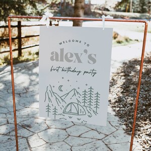 May include: A white sign with the text "Welcome to Alex's first birthday party" and a green illustration of a mountain range with a tent and stars.