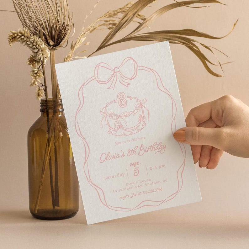 May include: A light pink birthday invitation with a bow and cake illustration. The invitation reads "Olivia's 8th Birthday" with event details. The invitation is held in front of a neutral background with a brown glass vase and dried plants.