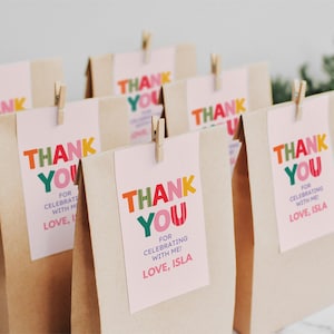 May include: A group of brown paper bags, each featuring a light pink rectangular tag attached with a wooden clothespin. The tags display the text THANK YOU FOR CELEBRATING WITH ME! LOVE, ISLA in colorful, bold, multi-colored block lettering.