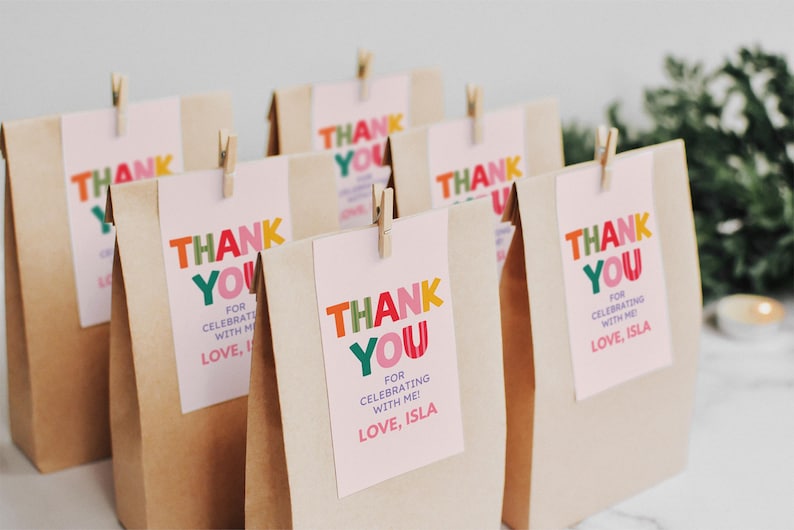 May include: A group of brown paper bags, each featuring a light pink rectangular tag attached with a wooden clothespin. The tags display the text THANK YOU FOR CELEBRATING WITH ME! LOVE, ISLA in colorful, bold, multi-colored block lettering.