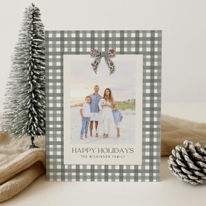 May include: A holiday card with a family photo, framed by a gray and white gingham pattern. The card features a decorative bow and the text "HAPPY HOLIDAYS" with a family name. A small Christmas tree and pinecone are in the background.
