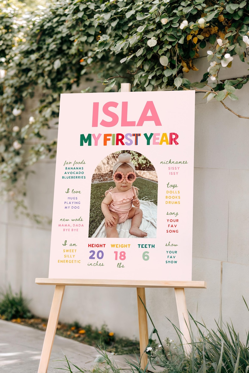 May include: A pink sign on a wooden easel displays the text "ISLA MY FIRST YEAR." It includes a photo of a baby wearing sunglasses, and lists facts such as height (20 inches), weight (18 lbs), and favorite foods. The sign is outdoors.