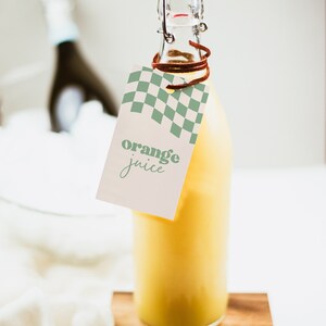 May include: A clear glass bottle filled with bright yellow orange juice, featuring a swing-top stopper with a red seal. A rectangular paper tag hangs from the neck by a brown cord, displaying a green checkered pattern and the text orange juice.