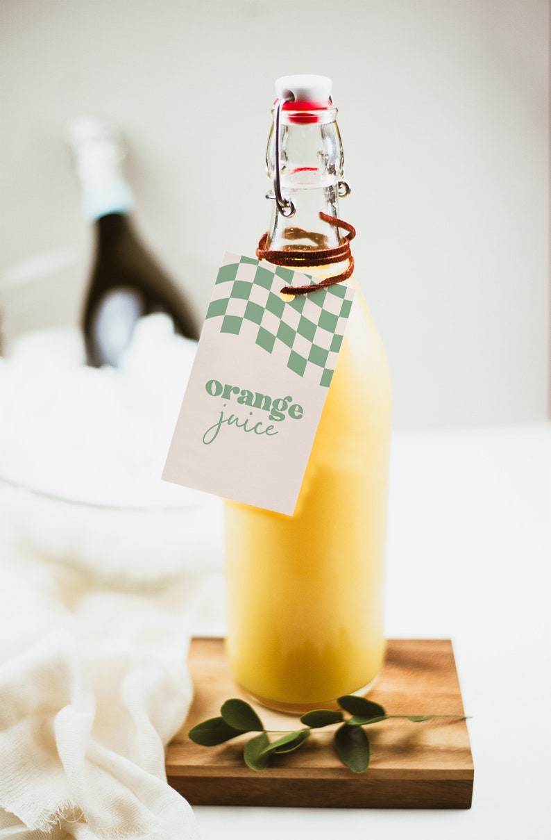 May include: A clear glass bottle filled with bright yellow orange juice, featuring a swing-top stopper with a red seal. A rectangular paper tag hangs from the neck by a brown cord, displaying a green checkered pattern and the text orange juice.