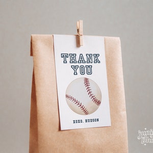 Peut inclure: Une carte de remerciement blanche avec du texte noir qui dit "THANK YOU" et un graphique de baseball. La carte est attach&eacute;e &agrave; un sac en papier brun avec une pince &agrave; linge en bois. Le texte au bas de la carte dit "XOXO. HUDSON".