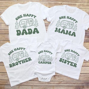 Puede incluir: Camisetas blancas con texto y gráficos verdes. Las camisetas dicen "ONE HAPPY DADA", "ONE HAPPY MAMA", "ONE HAPPY BROTHER", "ONE HAPPY CAMPER" y "ONE HAPPY SISTER". Cada camiseta presenta un gráfico de caravana.