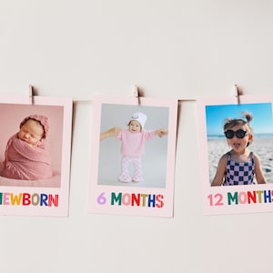May include: Three rectangular photo prints hanging on a string, each featuring a child at different ages. The prints are labeled with colorful text: NEWBORN, 6 MONTHS, and 12 MONTHS. The newborn is wrapped in pink, the 6-month-old wears a pink outfit, and the 12-month-old wears a checkered swimsuit.