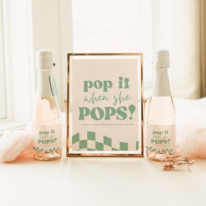 May include: Two glass bottles with custom labels featuring sage green text that reads pop it when she POPS! and a checkered pattern. Between the bottles stands a gold-framed sign with the same text and a sage green checkered base design.