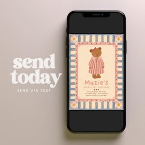 May include: A smartphone displaying a digital birthday invitation featuring a brown teddy bear in a pink striped dress. The invitation has a cream background, a blue and white striped border with pink hearts, and text reading Mazie's Beary First Birthday.