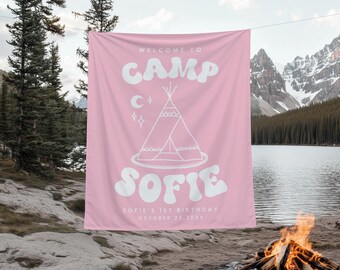 Puede incluir: Bandera de tela rosa con texto blanco: "WELCOME TO CAMP SOFIE". Presenta una ilustración de tipi, luna y estrellas. Incluye "SOFIE'S 1ST BIRTHDAY OCTOBER 23, 20XX". La bandera cuelga al aire libre cerca de un lago y montañas.