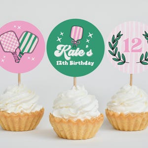 May include: Three cupcakes with white frosting, each topped with a decorative circle. One pink circle features pickleball paddles, another green circle says "Kate's 12th Birthday", and the last has a pink "12" with a green wreath.