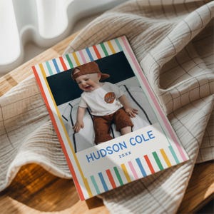 Puede incluir: Una impresi&oacute;n fotogr&aacute;fica rectangular que muestra a un ni&ntilde;o con gorra marr&oacute;n, camiseta blanca y pantalones cortos marrones. La impresi&oacute;n tiene un borde de rayas verticales de colores y el texto azul "HUDSON COLE" y "20XX" sobre fondo blanco.