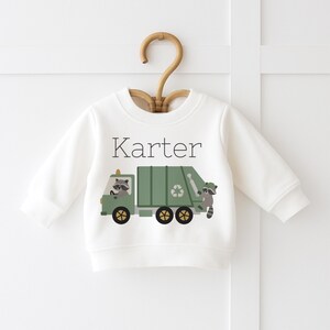 May include: A white baby sweatshirt featuring a green garbage truck design with raccoons and a recycling symbol. The name "Karter" is printed above the truck. The sweatshirt is displayed on a wooden hanger.